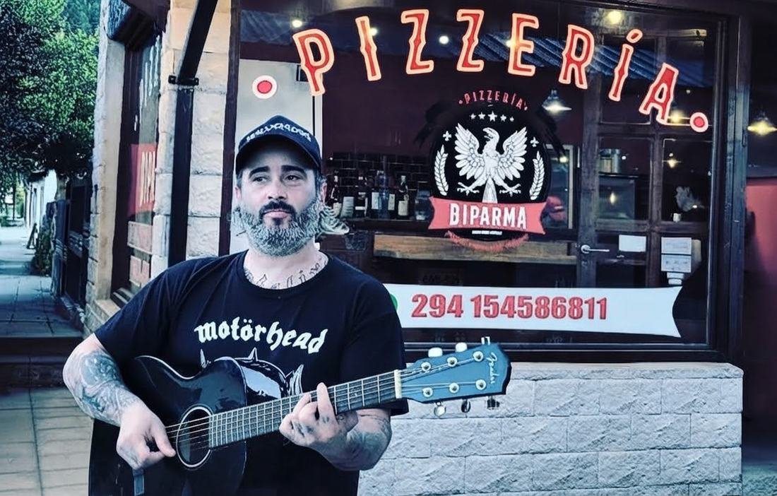 Un músico con guitarra acústica en la puerta de Diparma, bajo el cartel de neón de la pizzería